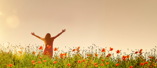 A woman in a peach-colored dress stands in a field of orange flowers. She has her arms thrown up wide in the air and her head thrown back.